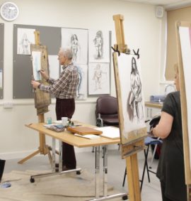 4 people in a classroom with easels drawing a live model