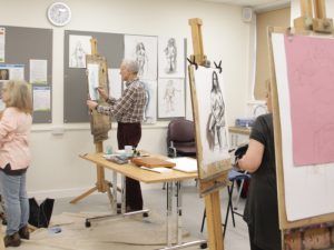 4 people in a classroom with easels drawing a live model