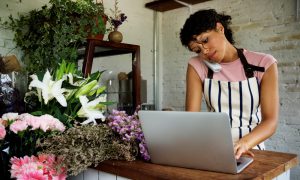 Woman wearing pink tshirt and stripy apron on telephone and typing at a laptop in a florist