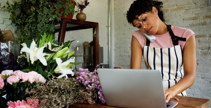 Woman wearing pink tshirt and stripy apron on telephone and typing at a laptop in a florist